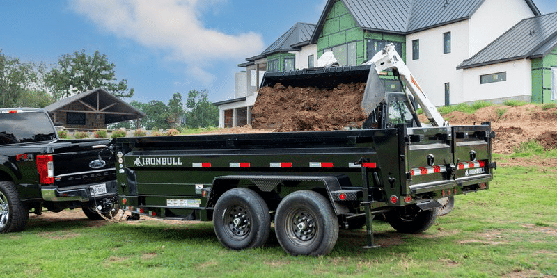 Iron Bull dump trailer hauling a skid steer behind a pickup truck at a job site near Broken Arrow, OK